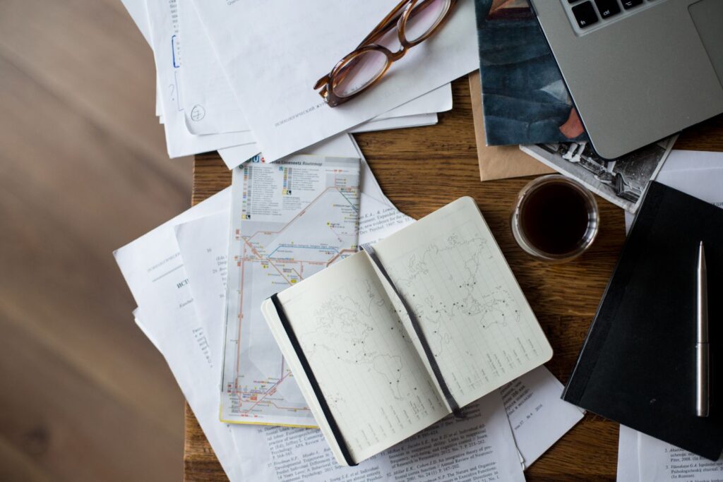 A desk with a laptop, maps, notes and a cup of coffee