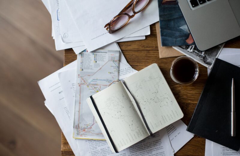 A desk with a laptop, maps, notes and a cup of coffee