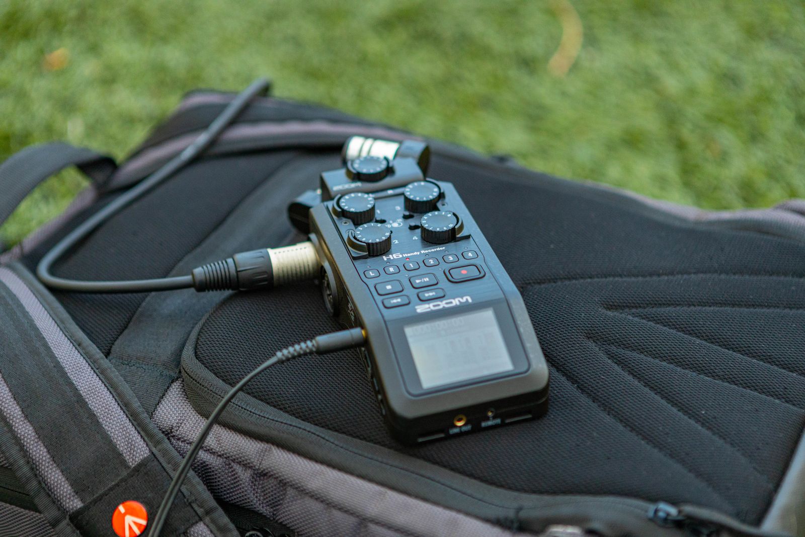 A portable audio recorder with cables lying on a backpack in the grass