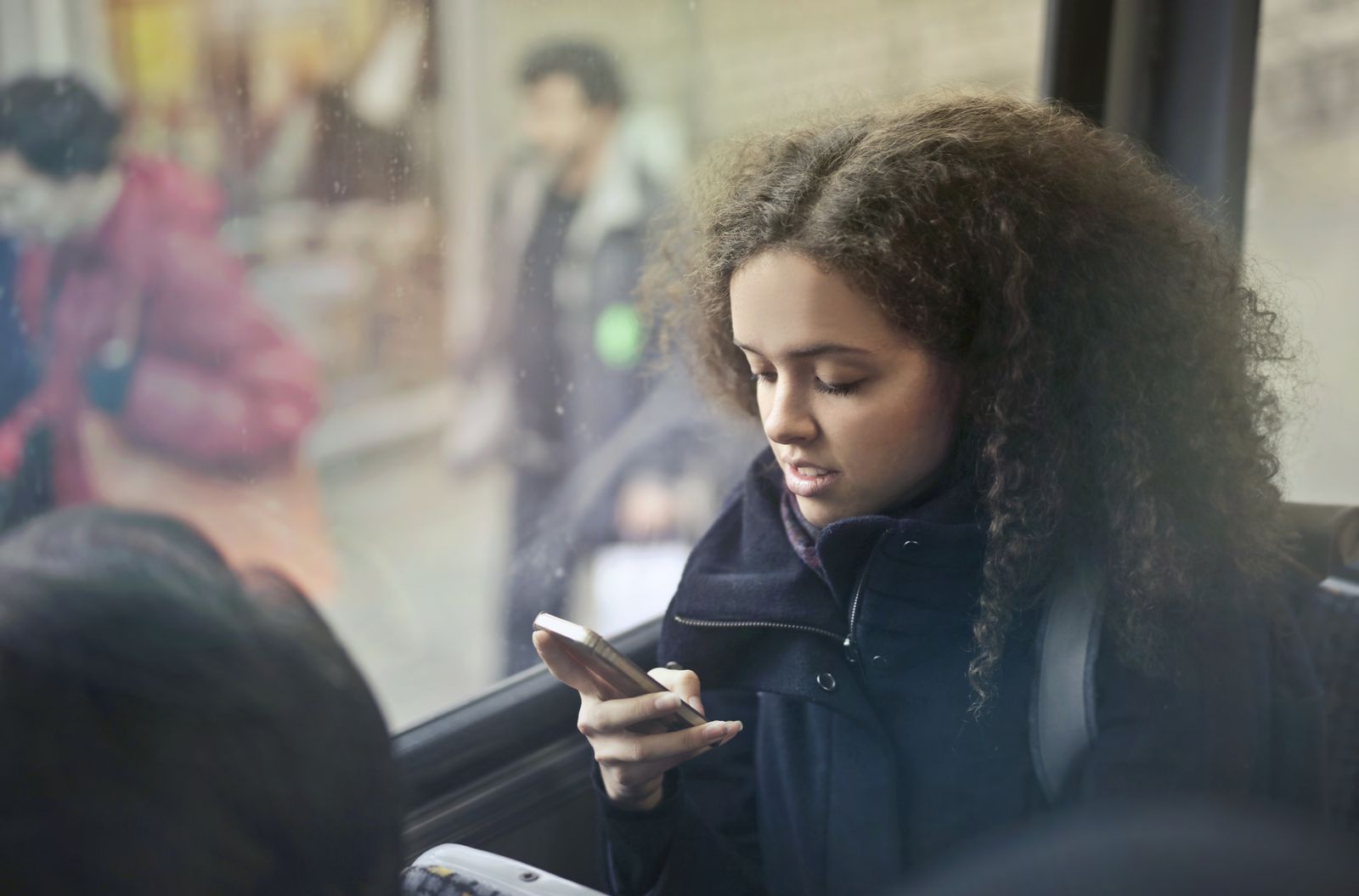 A young woman with curly hair reading her phone by the window of a bus