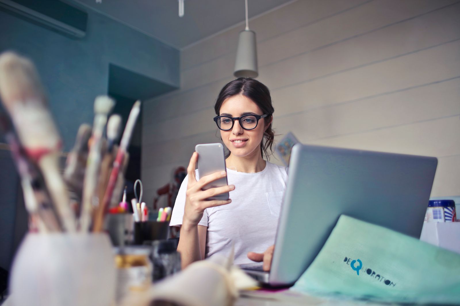 A woman at a desk in a studio checking her phone beside a laptop and art supplies