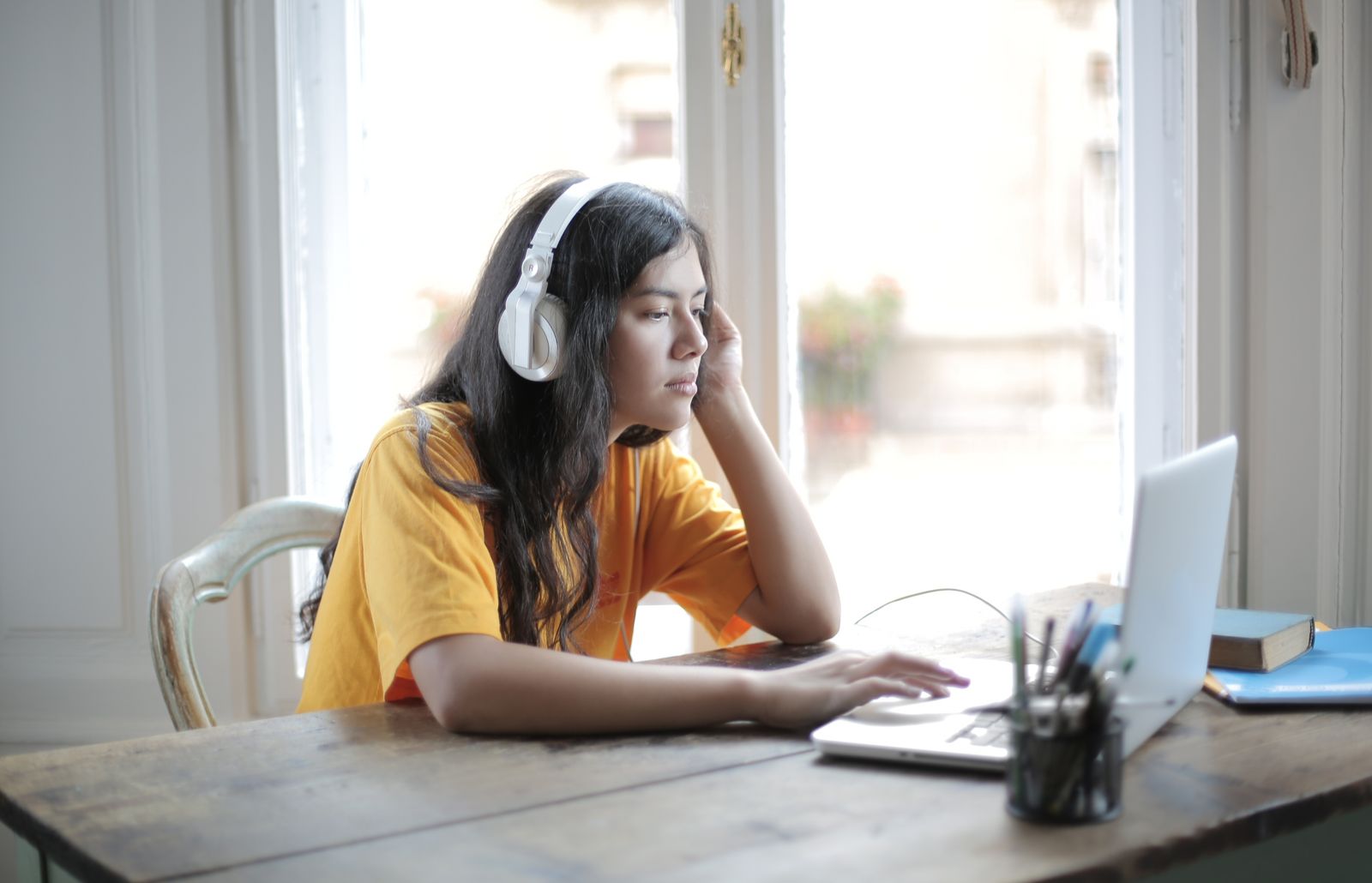 A woman in headphones listening at a laptop on a wooden table by the window