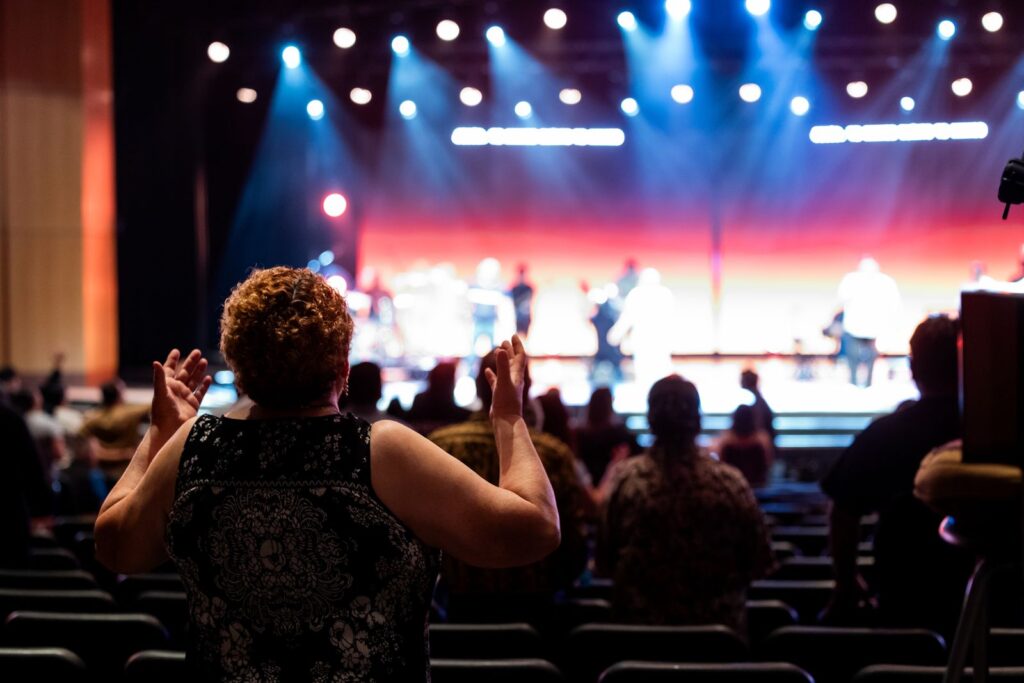 A concert audience clapping in front of a lit stage, seen from behind