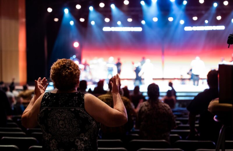 A concert audience clapping in front of a lit stage, seen from behind