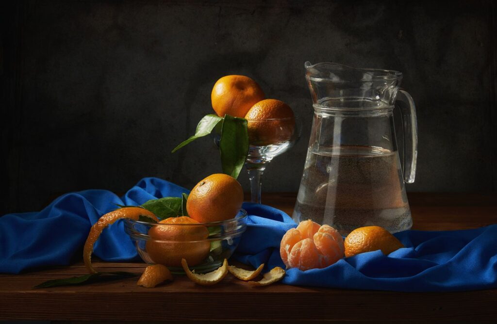 A still life of tangerines and a glass jug on a blue cloth