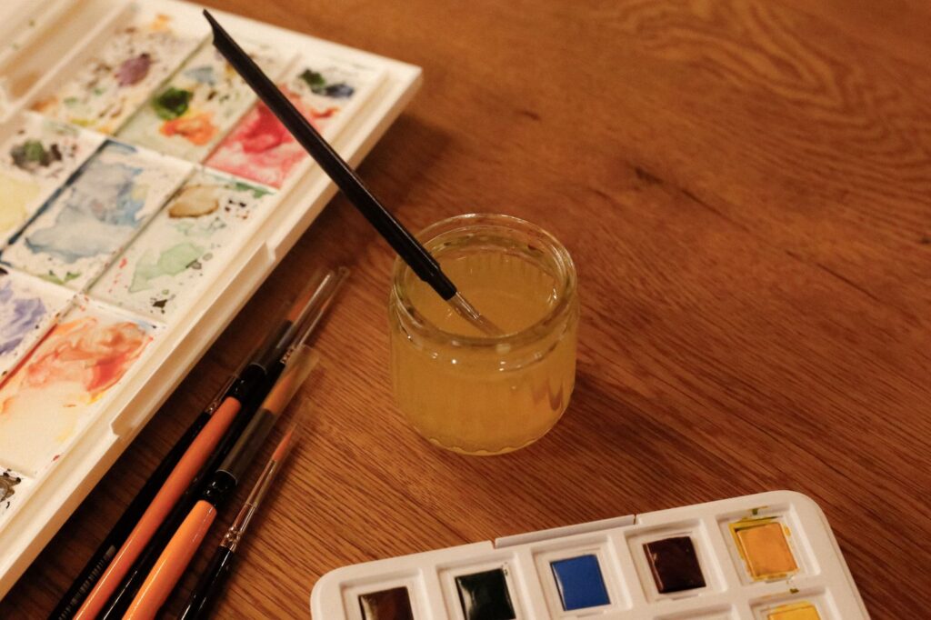 A watercolour paint box, brushes and a jar of water on a wooden table