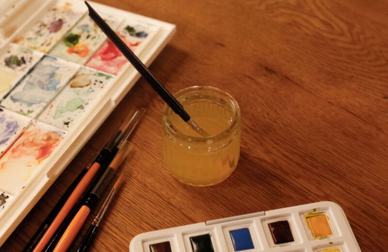 A watercolour paint box, brushes and a jar of water on a wooden table