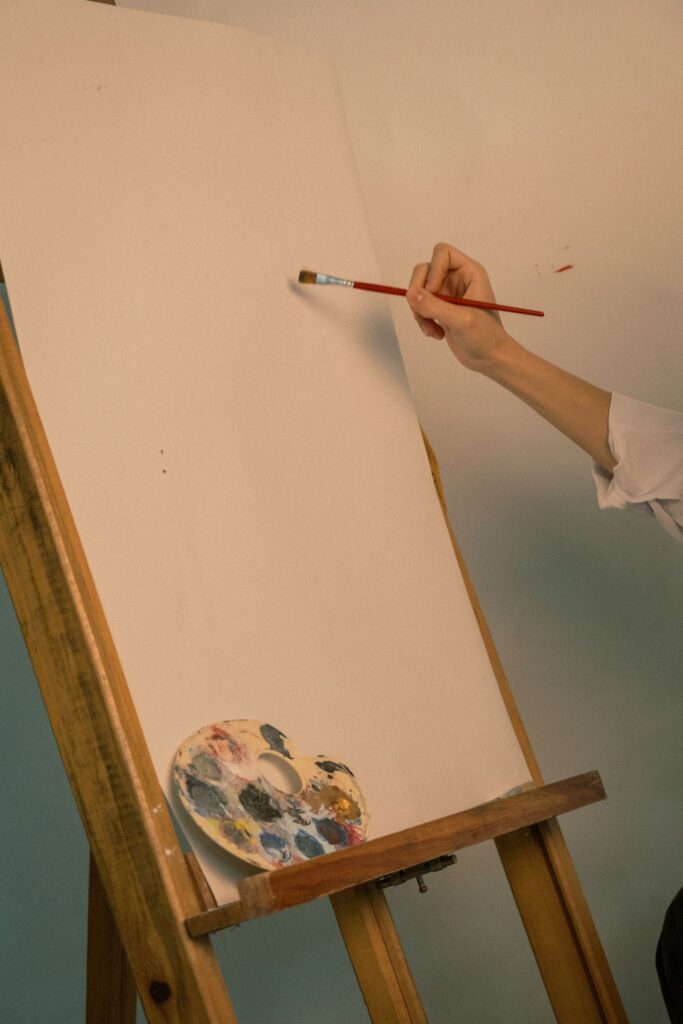 A hand with a fine brush starting a blank canvas on an easel