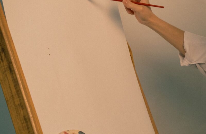 A hand with a fine brush starting a blank canvas on an easel