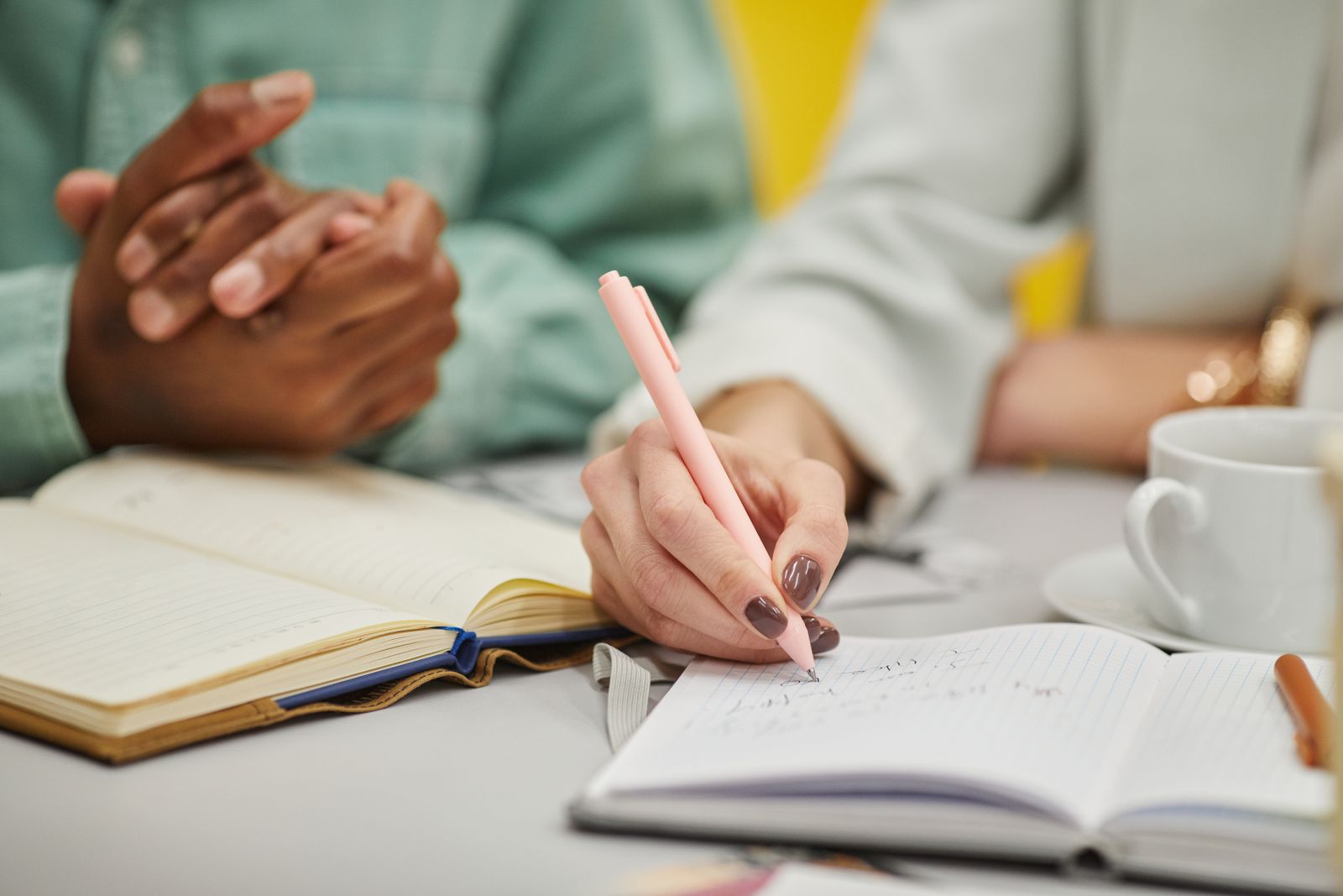 Hands writing in notebooks at a meeting table, a coffee cup beside them