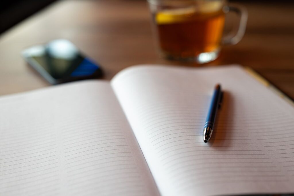 An open notebook with a pen, a phone and a cup of tea on a table