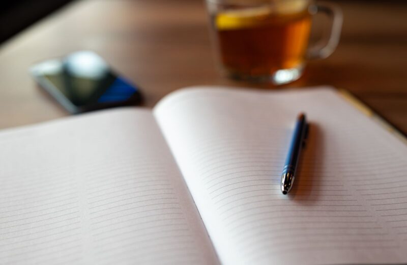 An open notebook with a pen, a phone and a cup of tea on a table