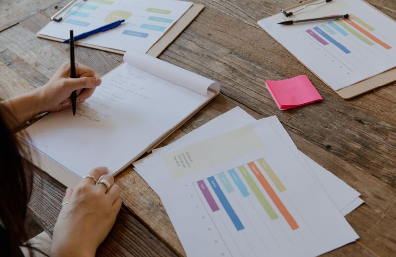 Hands writing notes beside printed bar charts on a wooden table