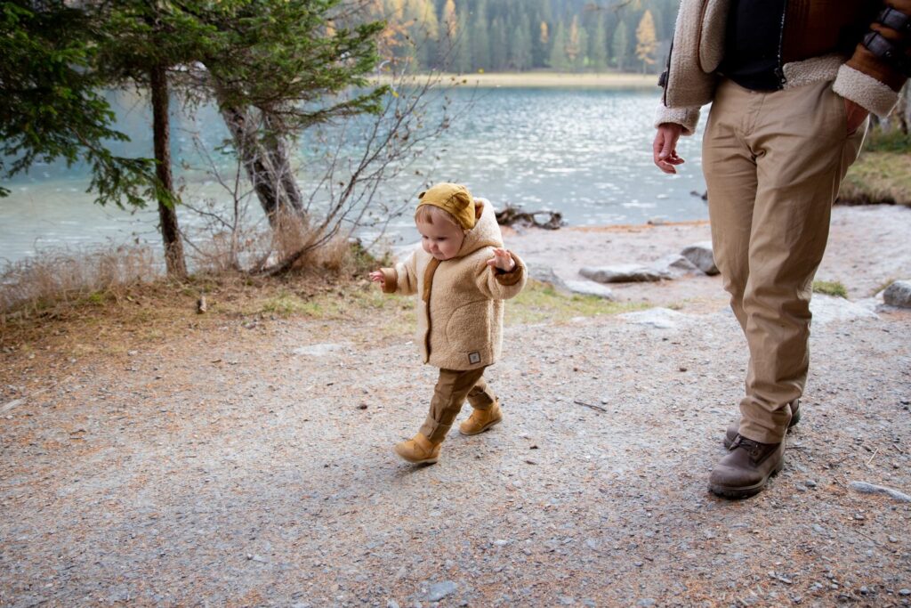 A toddler in a warm jacket and boots walking beside a lake with an adult