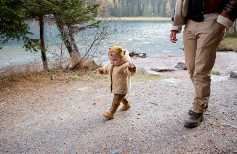 A toddler in a warm jacket and boots walking beside a lake with an adult