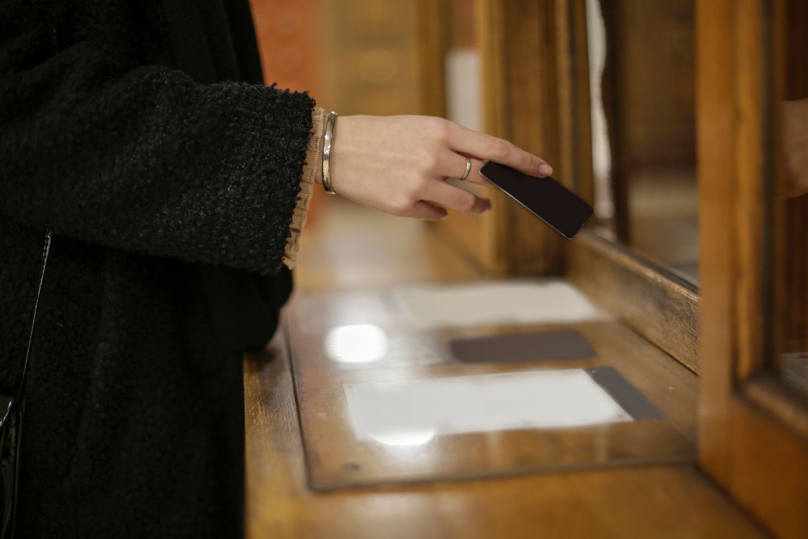 A customer's hand at a wooden bank counter window, about to slide a card under the glass