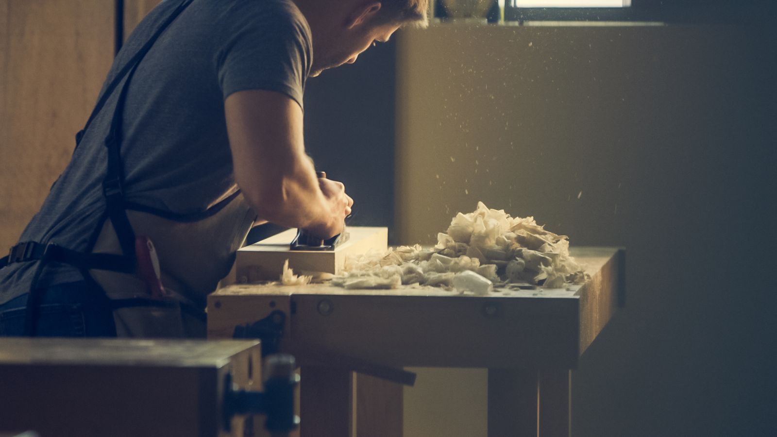 A craftsman shaping a piece of wood at his workshop bench, shavings around him