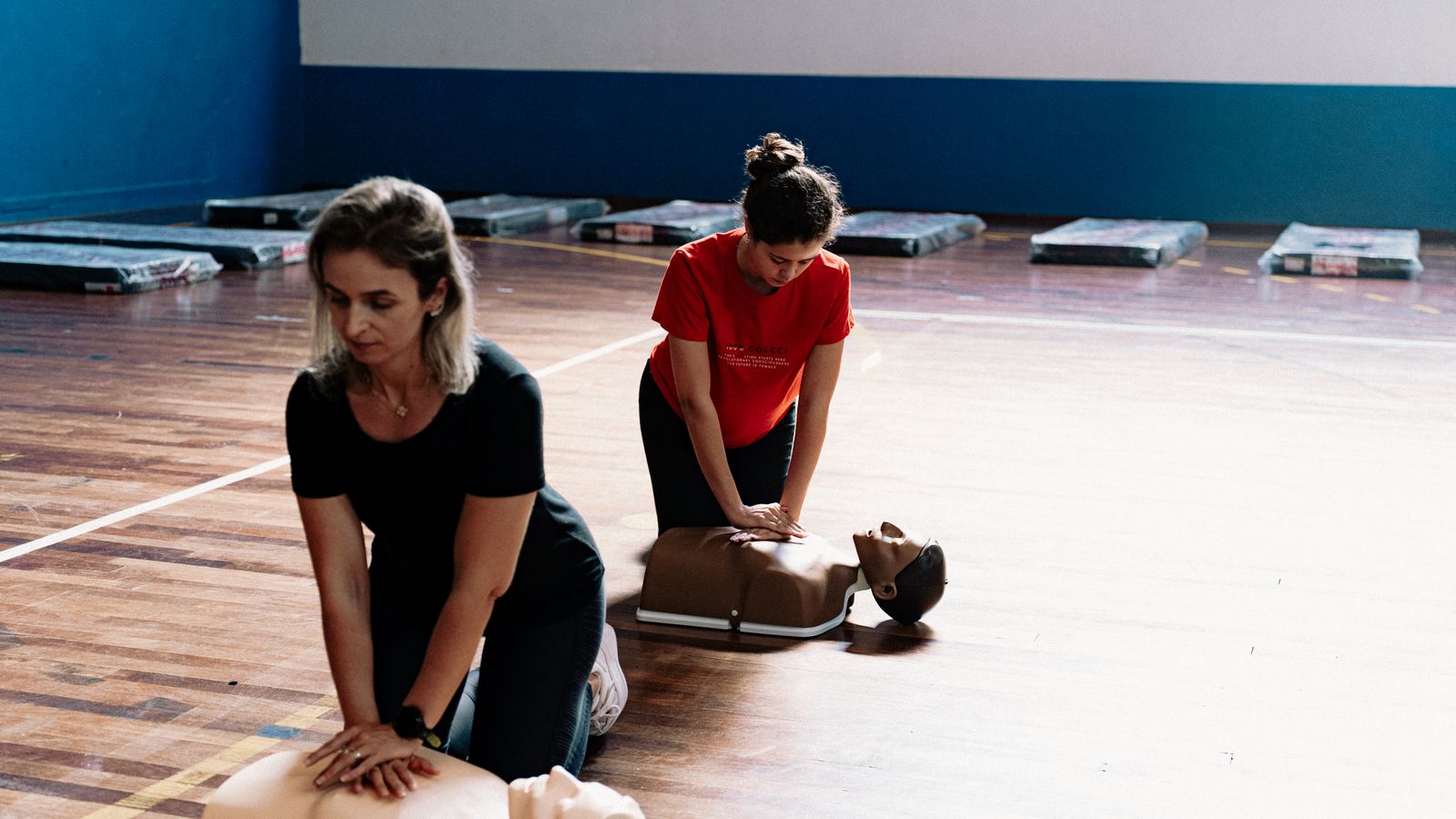 Two women practising chest compressions on manikins in a sports hall during a first-aid course