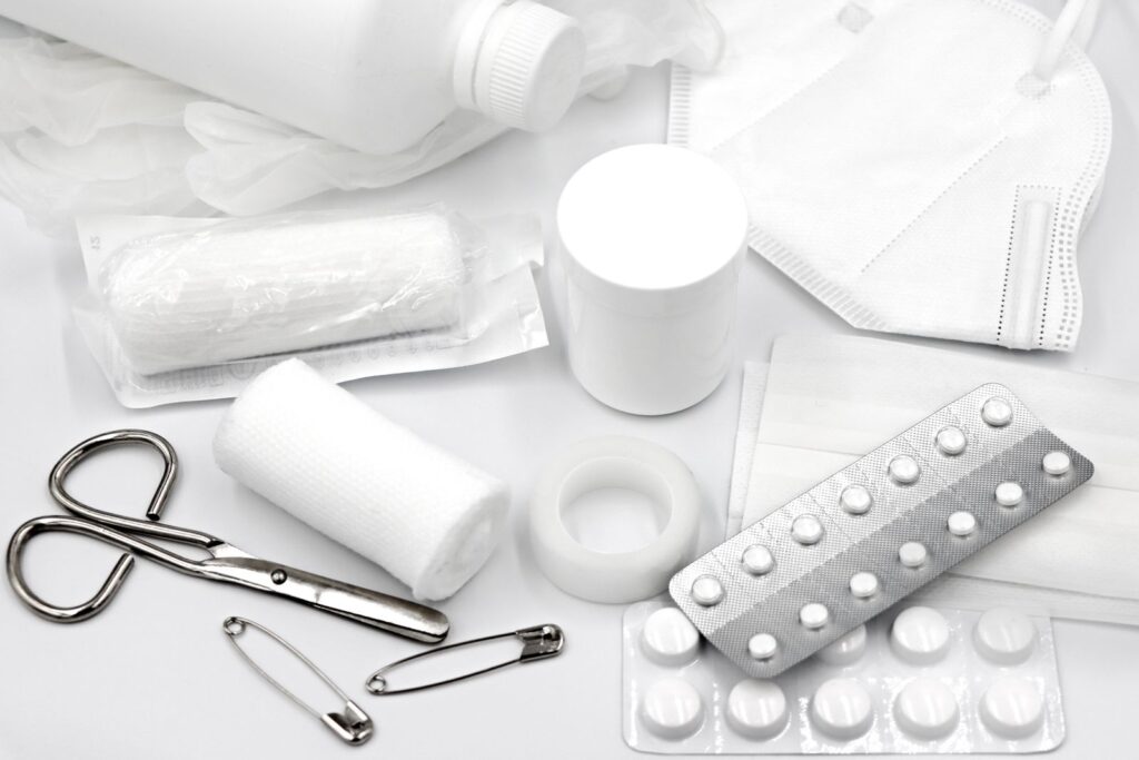 First-aid supplies laid out on a white table: bandages, tape, scissors, safety pins, a mask and blister packs