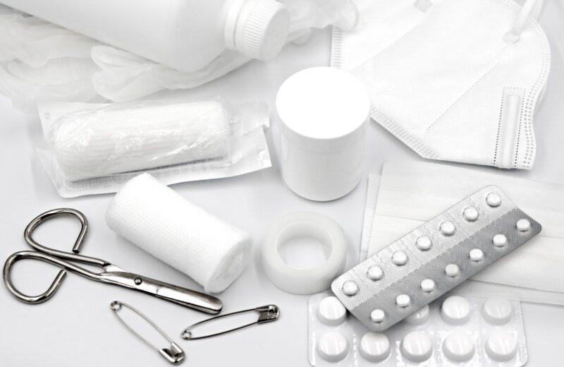 First-aid supplies laid out on a white table: bandages, tape, scissors, safety pins, a mask and blister packs