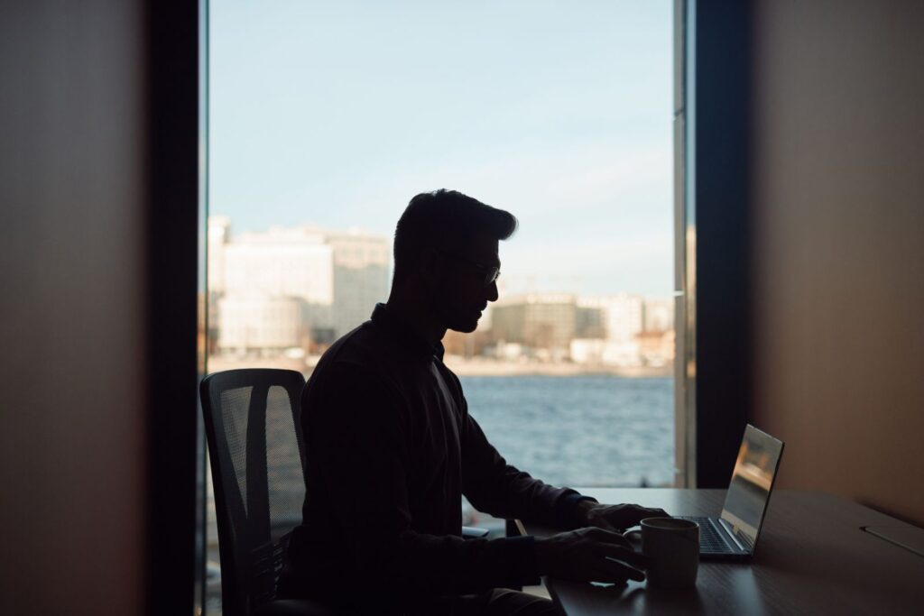 A person in silhouette working on a laptop by a large window