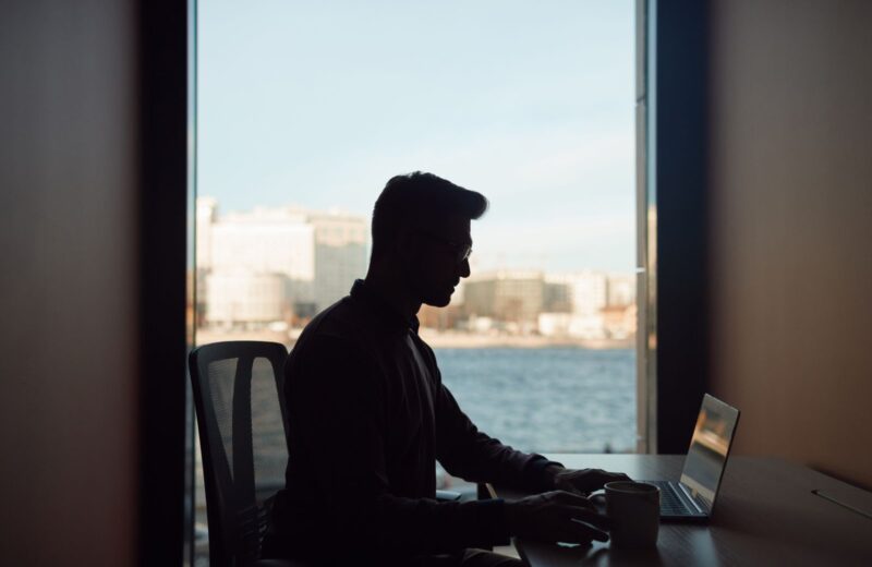 A person in silhouette working on a laptop by a large window
