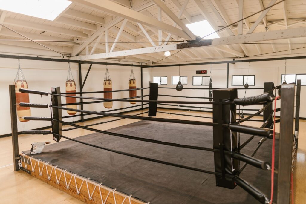An empty boxing ring under a high roof with heavy bags behind it