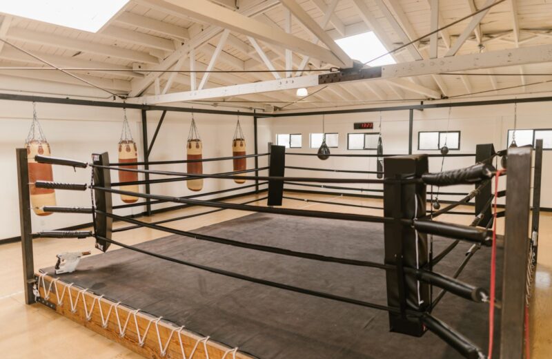 An empty boxing ring under a high roof with heavy bags behind it