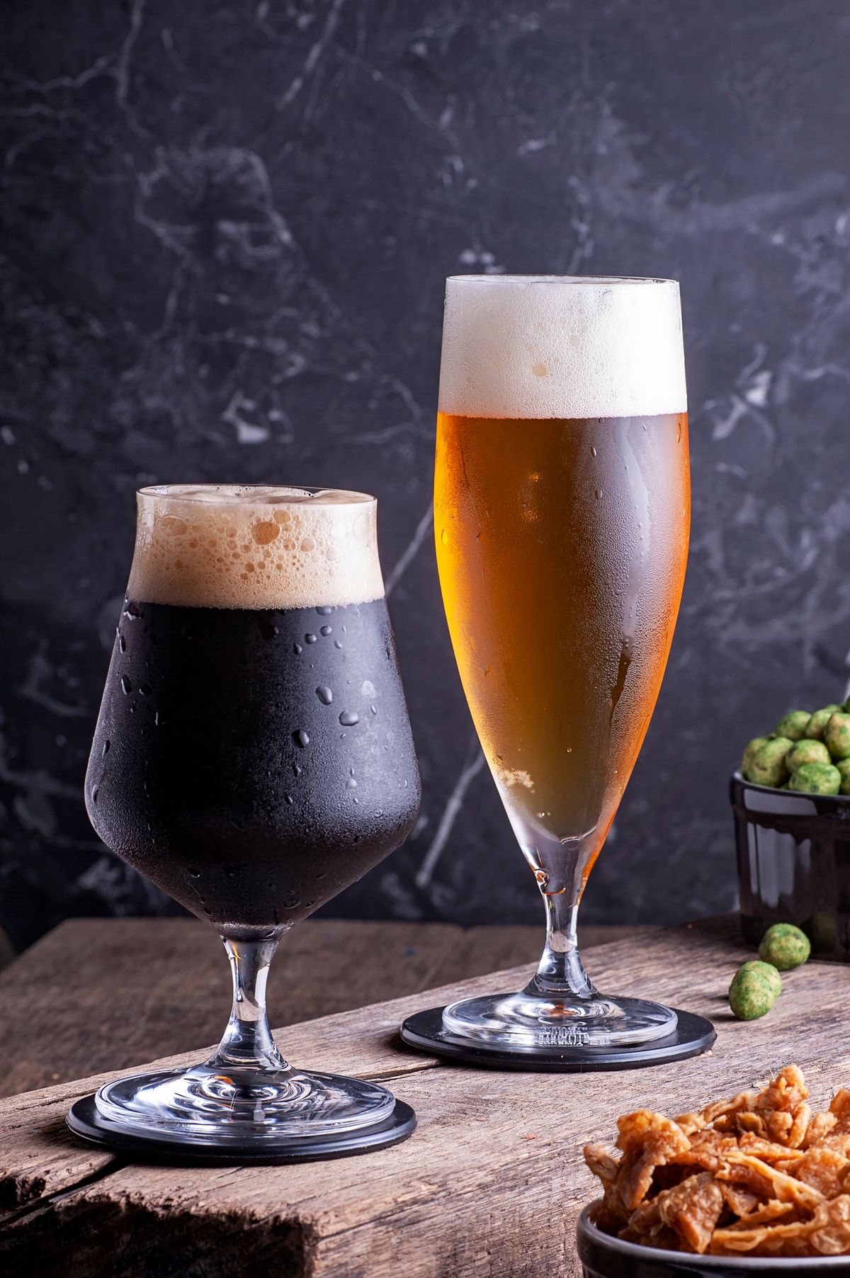 A glass of stout and a tall glass of golden beer on dark stone, with hops and pretzels beside them