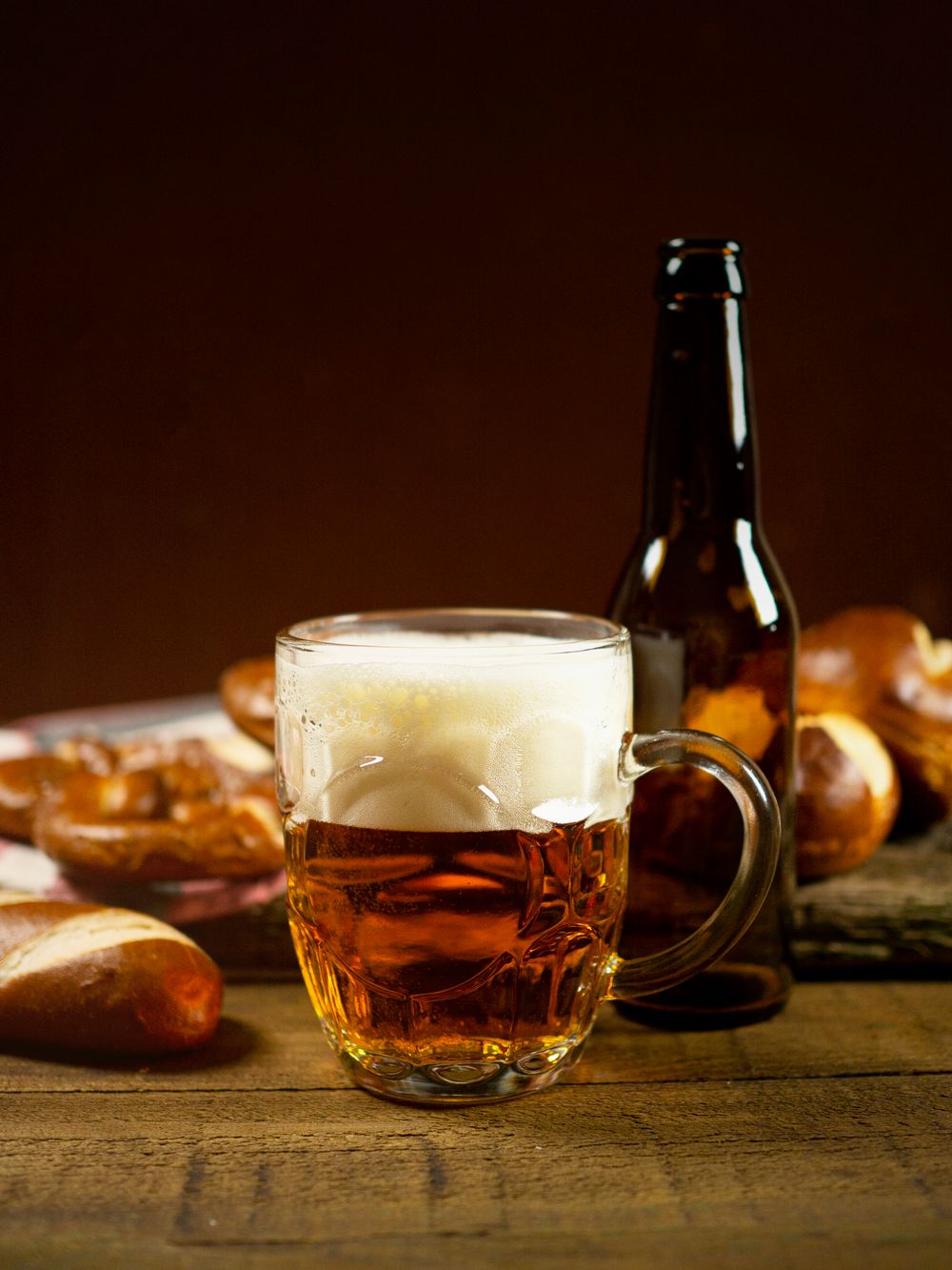 A glass mug of amber beer and a brown bottle on a wooden table with pretzels