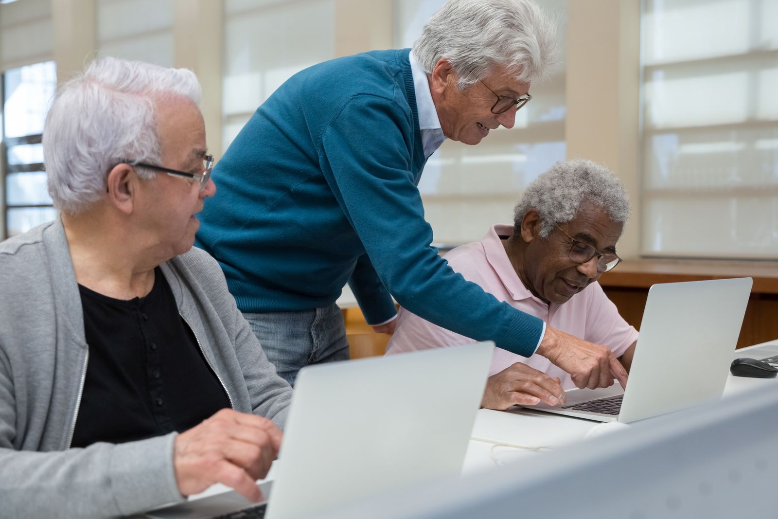 A teacher leaning over to help two older students working on laptops in a bright classroom