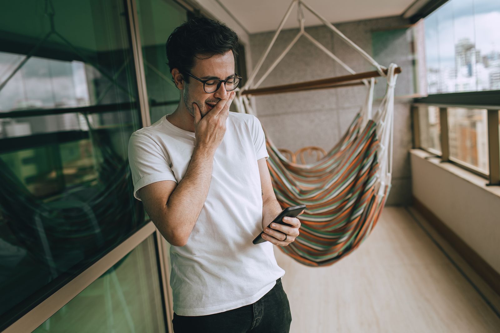 A man in glasses reading a message on his phone, his hand at his chin