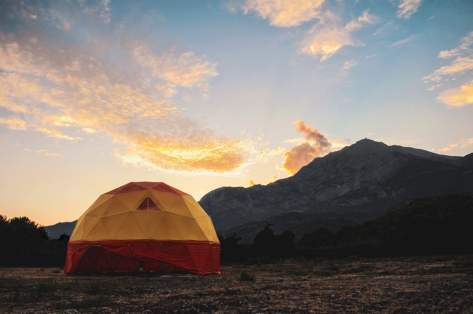 A red and yellow geodesic tent on open ground under a sunset sky, mountains behind