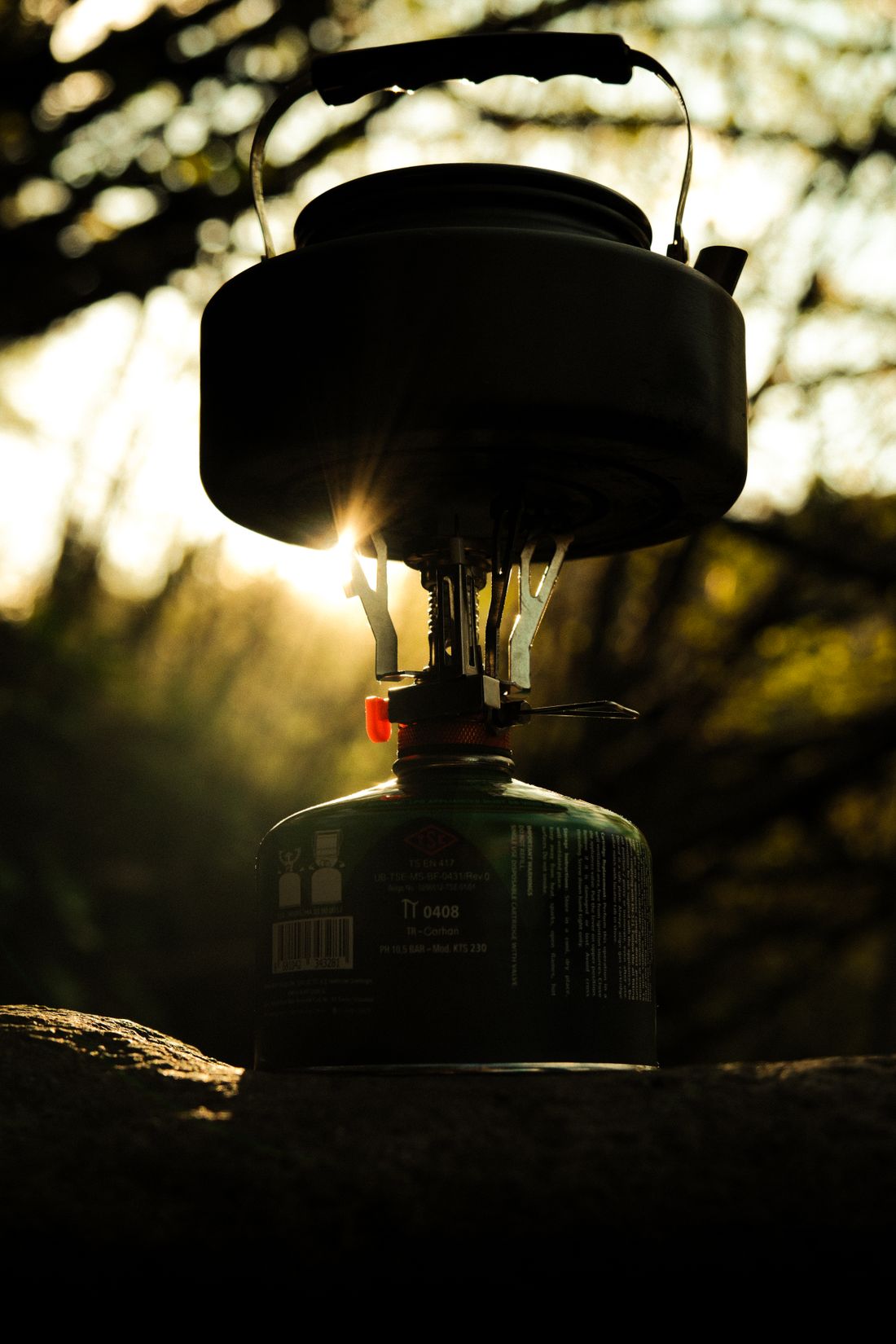 A small gas stove with a pot on top, backlit by the evening sun in long grass