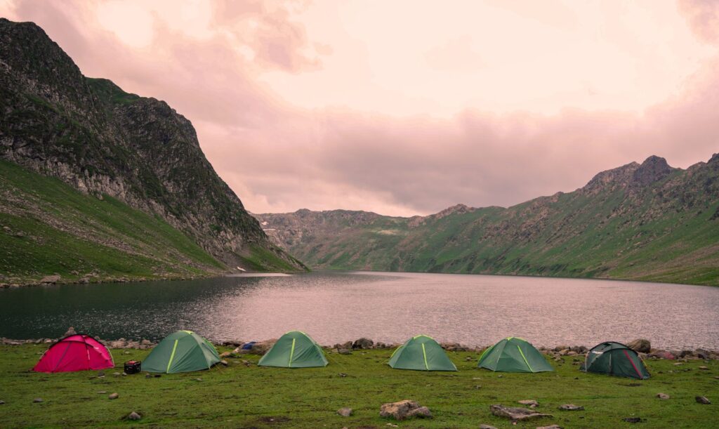 Small tents in a row on a green meadow by a mountain lake under a pink evening sky