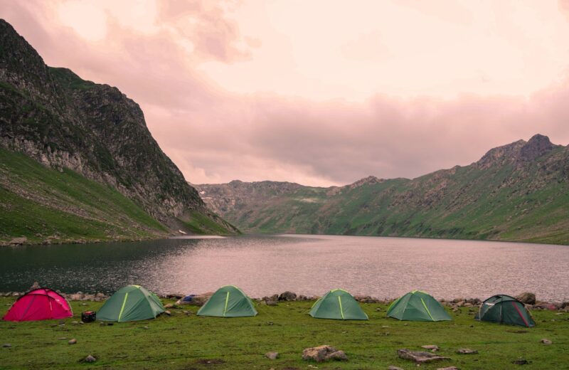Small tents in a row on a green meadow by a mountain lake under a pink evening sky
