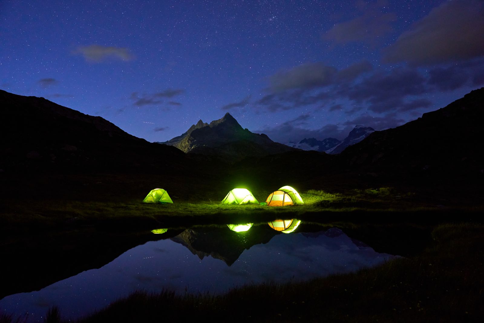 Three tents lit from inside, reflected in a mountain lake at night