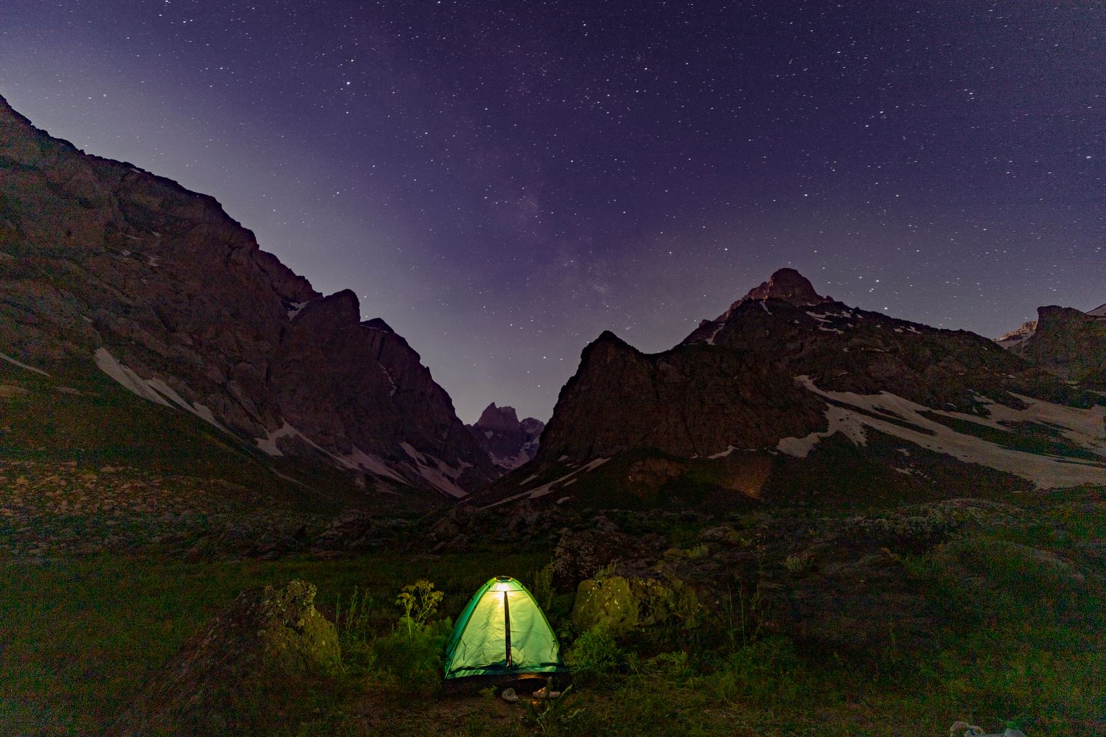 A small tent glowing green in a valley under a starry sky between dark mountains