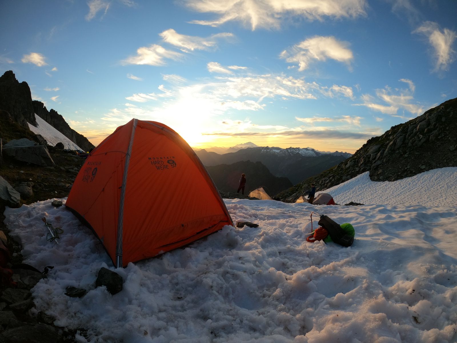 An orange dome tent pitched on snow below a mountain ridge, the sun low behind it