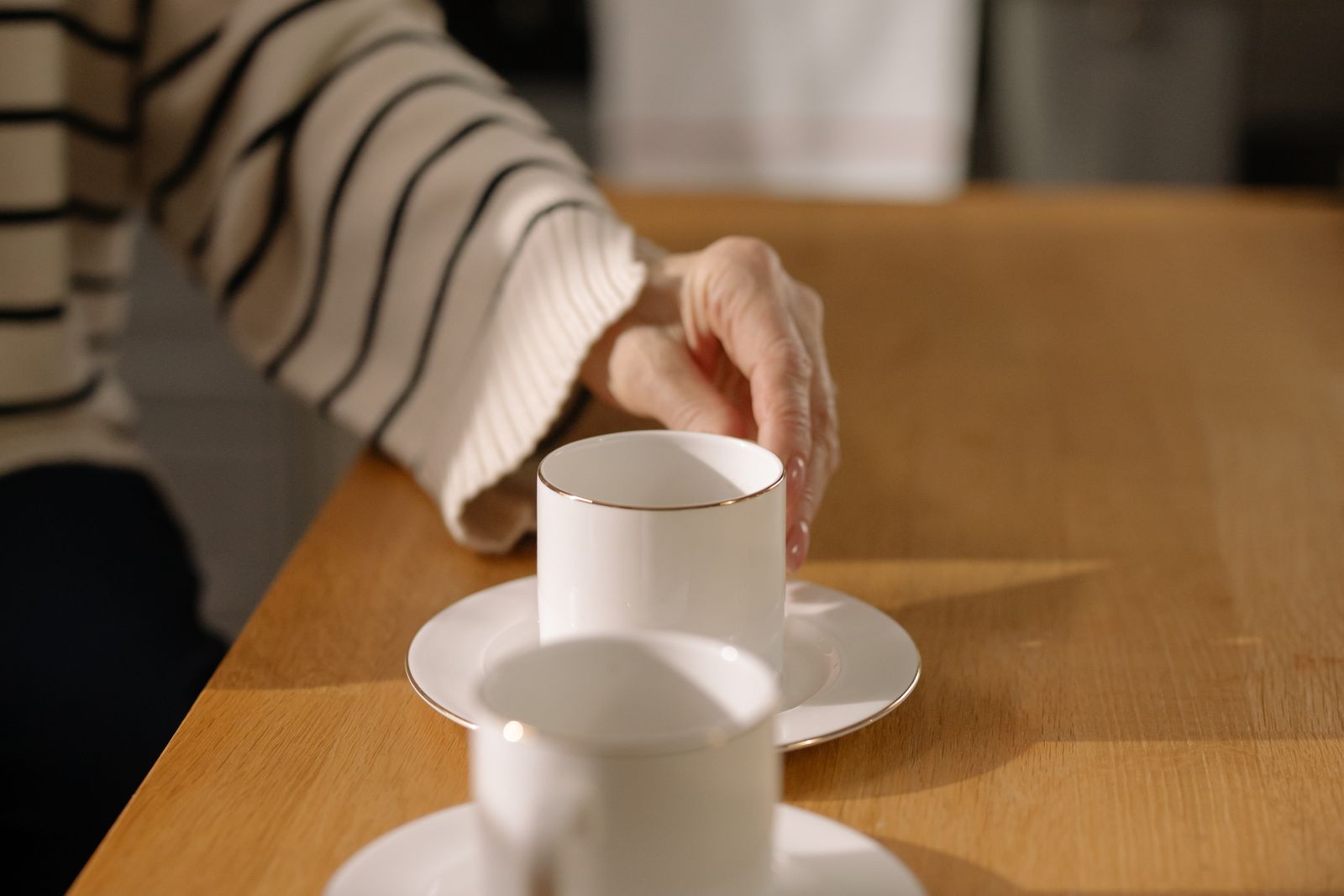 A hand reaching for a white cup and saucer on a kitchen table