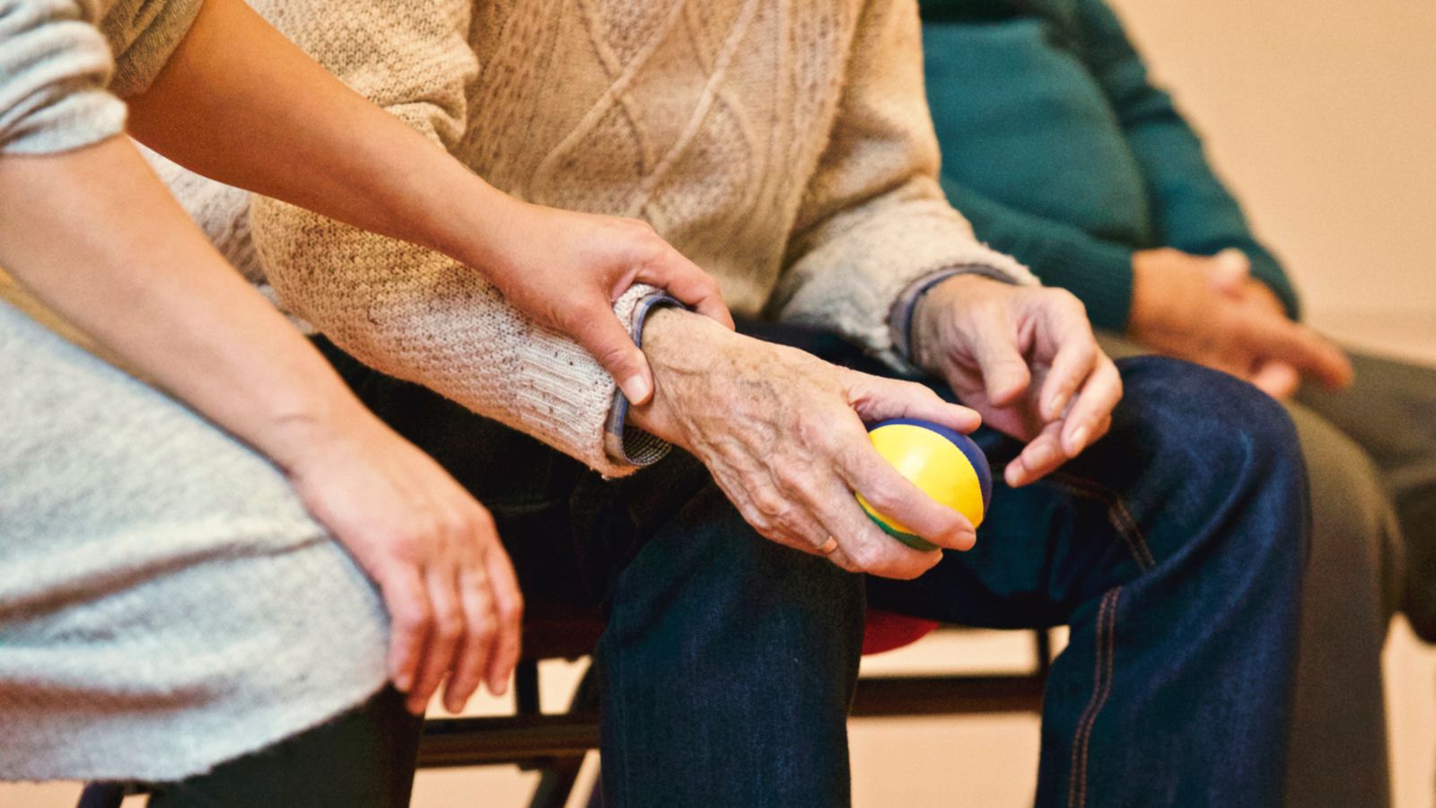 A carer's hand resting on an older man's hand as he holds a soft ball, faces out of view