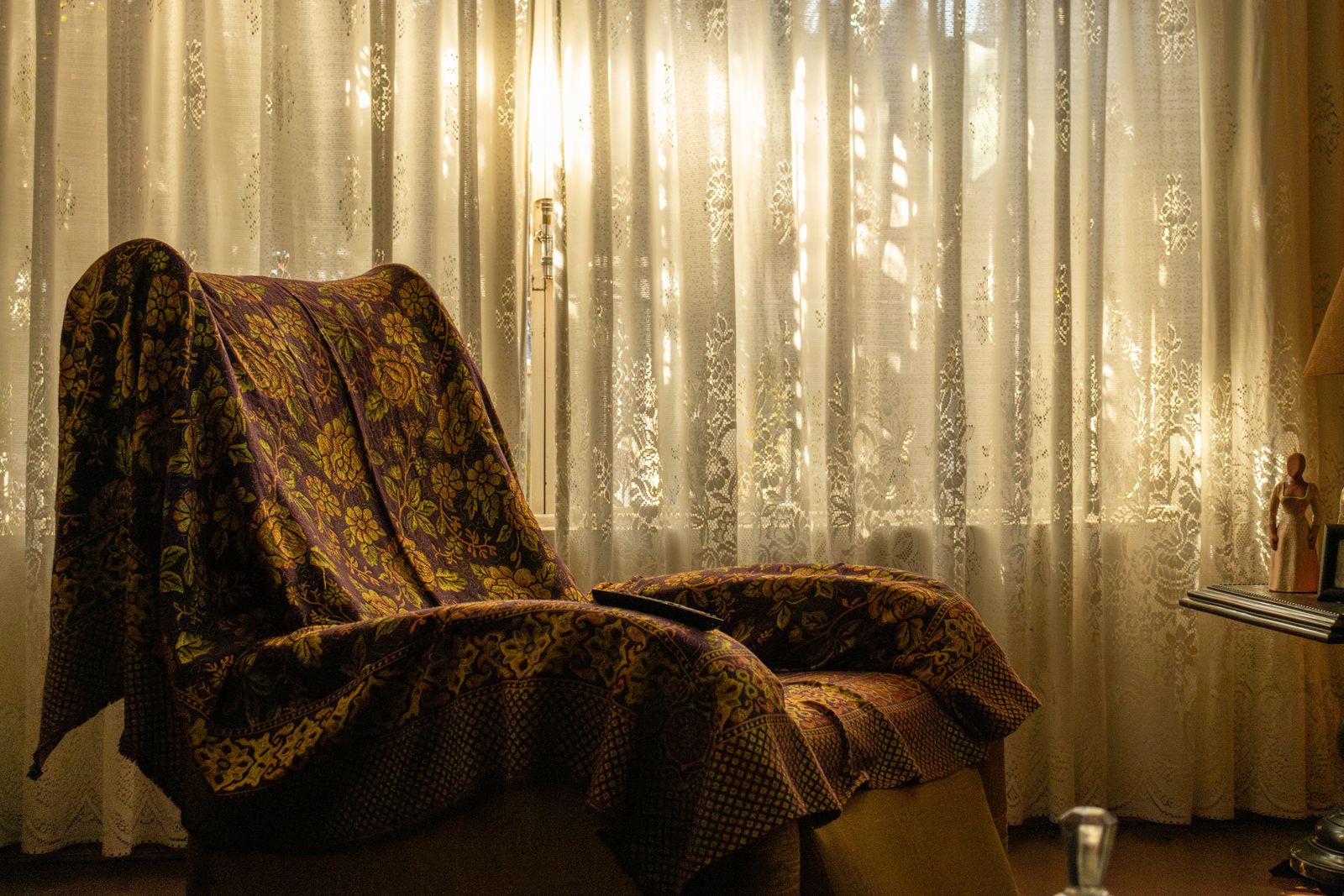 An armchair with a floral blanket beside a curtained window in soft light