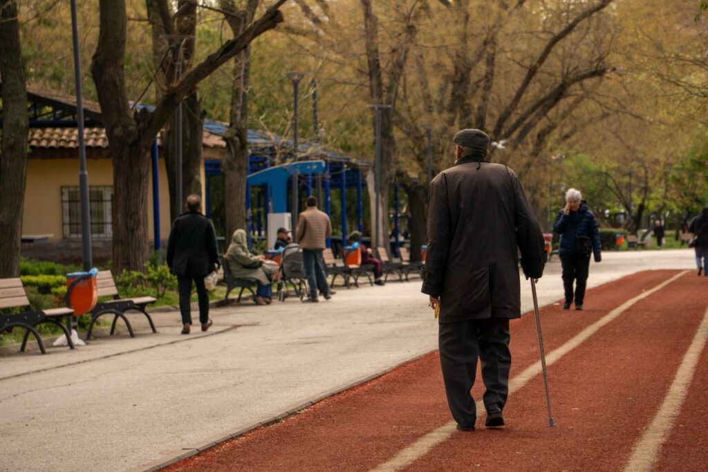 An older man with a walking cane on a tree-lined park path, seen from behind