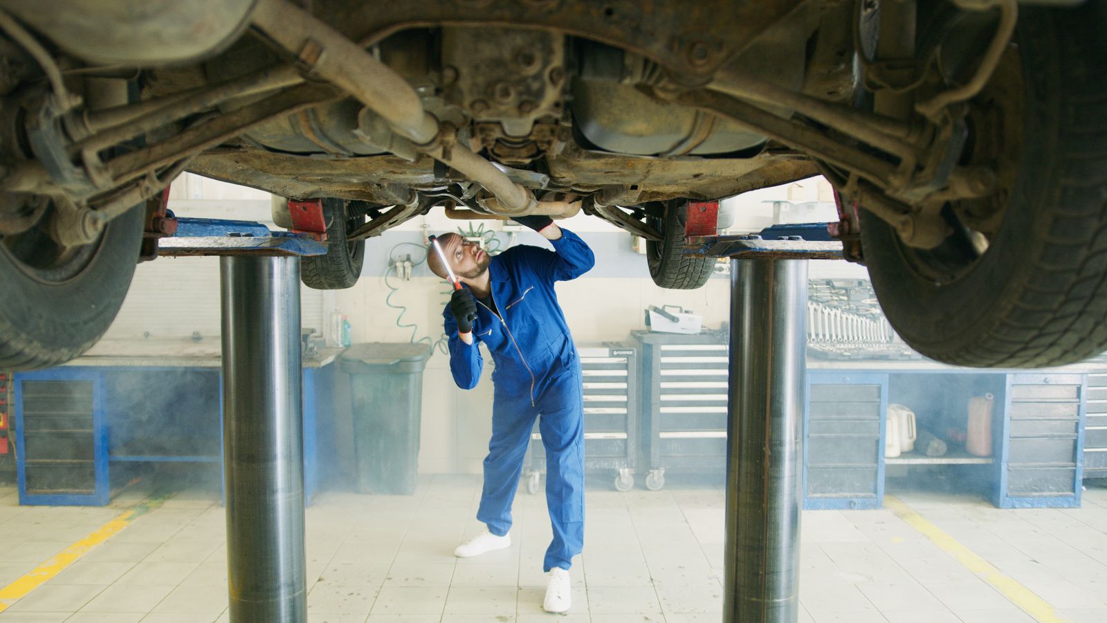 A technician in blue overalls standing under a car raised on a two-post lift