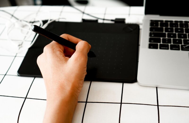 A hand with a pen tablet beside a laptop on a white desk