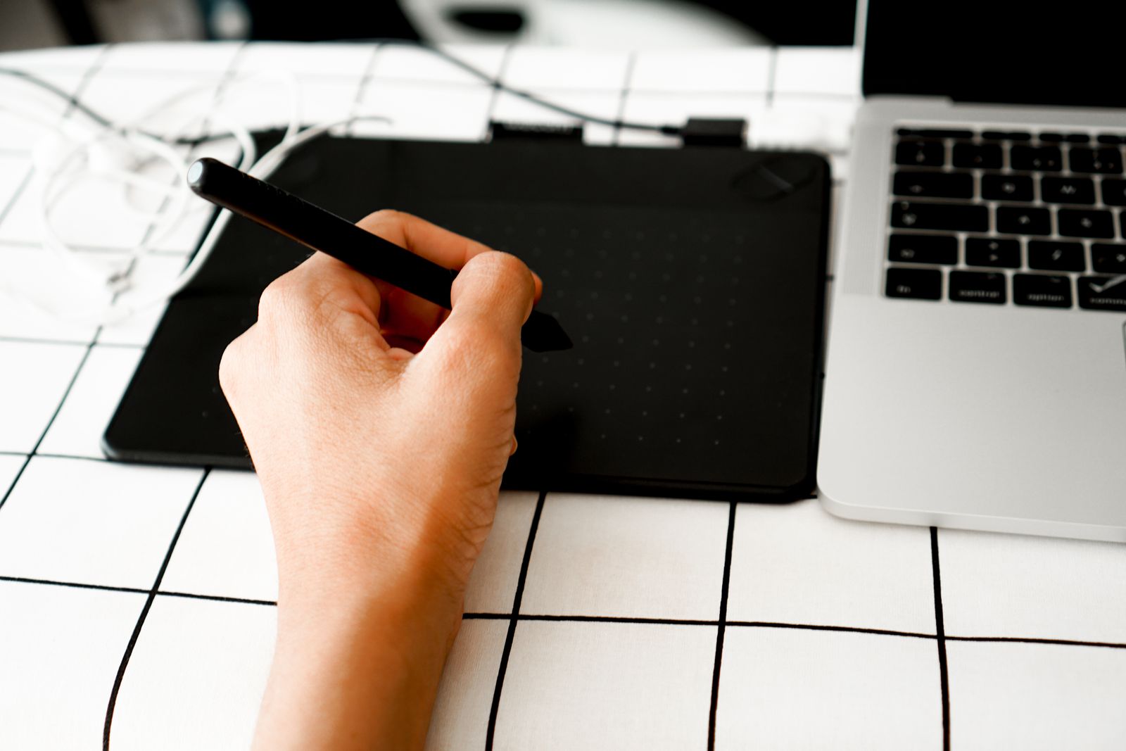 A hand with a pen tablet beside a laptop on a white desk