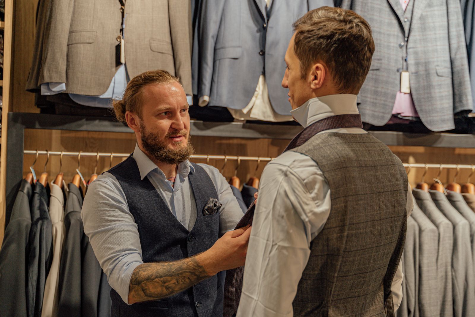 A shop assistant in a waistcoat adjusting a customer's tie in front of a wall of jackets