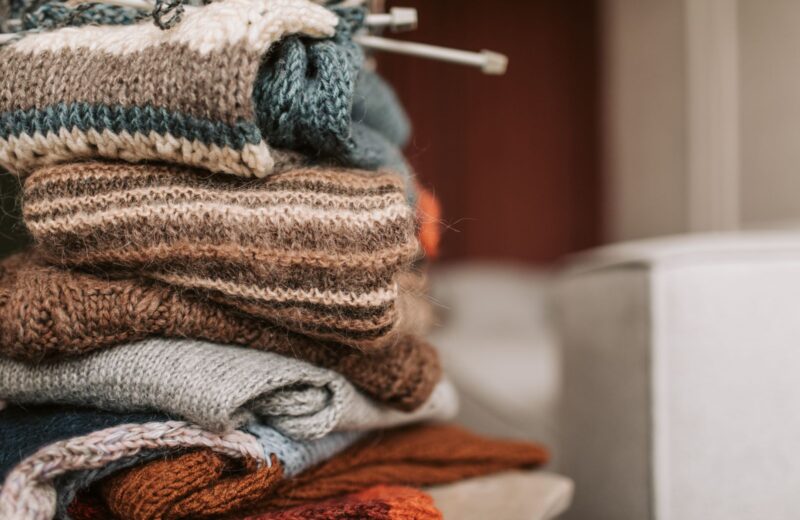 A tall stack of hand-knitted jumpers in brown, grey and rust with knitting needles on top