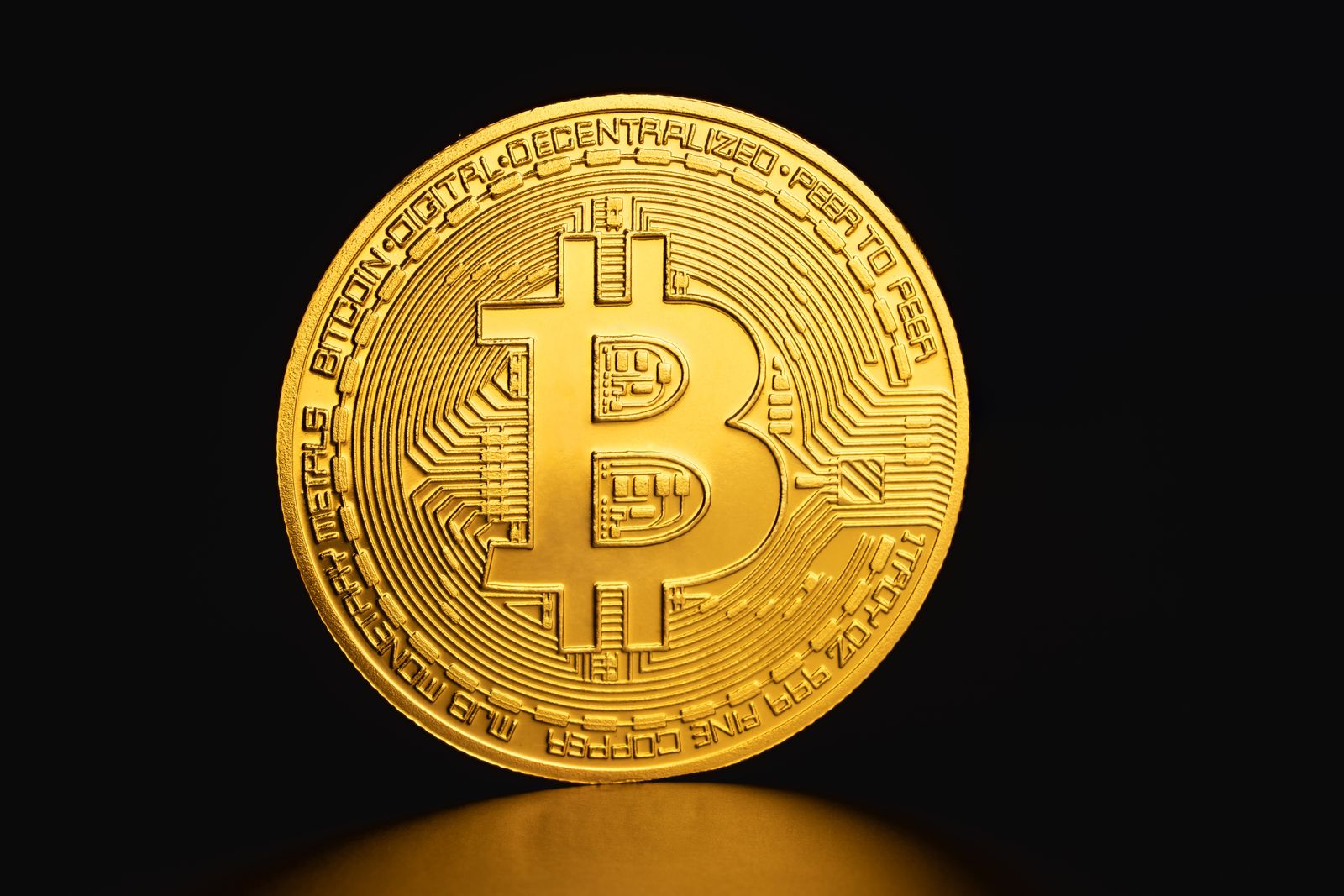 A single gold-coloured Bitcoin coin standing on a dark surface, lit from above