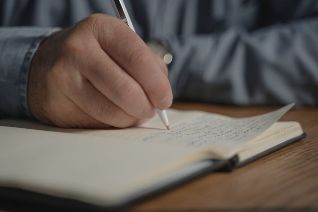 A hand writing with a pen in an open notebook on a wooden desk